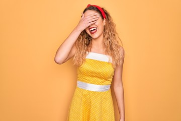 Beautiful blonde pin-up woman with blue eyes wearing diadem standing over yellow background smiling and laughing with hand on face covering eyes for surprise. Blind concept.
