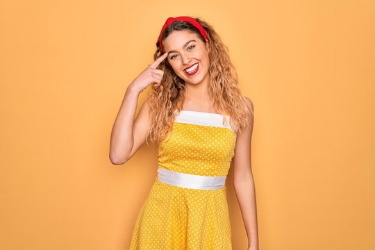 Beautiful blonde pin-up woman with blue eyes wearing diadem standing over yellow background Smiling pointing to head with one finger, great idea or thought, good memory
