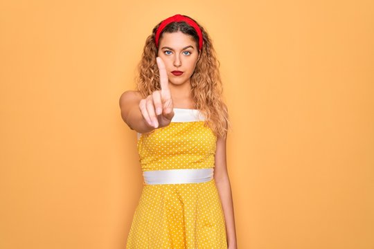 Beautiful blonde pin-up woman with blue eyes wearing diadem standing over yellow background Pointing with finger up and angry expression, showing no gesture