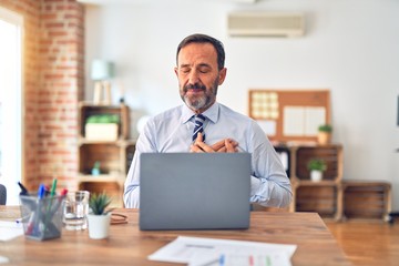 Middle age handsome businessman wearing tie sitting using laptop at the office smiling with hands on chest with closed eyes and grateful gesture on face. Health concept.