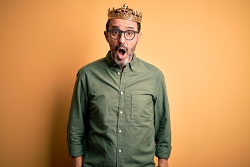 Middle age hoary man wearing king crown standing over isolated yellow background afraid and shocked with surprise expression, fear and excited face.