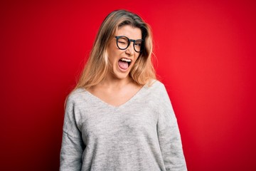 Young beautiful blonde woman wearing sweater and glasses over isolated red background winking...