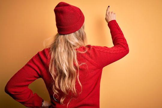 Young beautiful blonde woman wearing casual sweater and wool cap over white background Posing backwards pointing ahead with finger hand