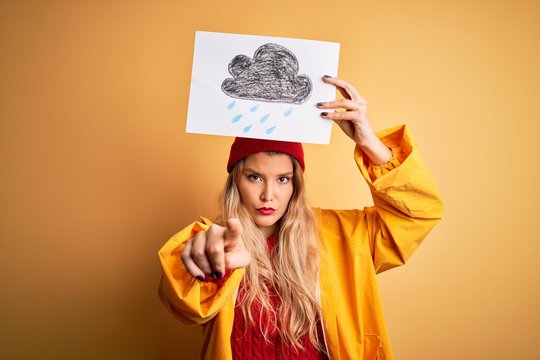 Young beautiful blonde woman wearing raincoat and wool cap holding banner with cloud image pointing with finger to the camera and to you, hand sign, positive and confident gesture from the front