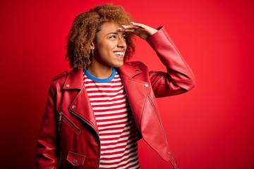 Young beautiful African American afro woman with curly hair wearing casual red jacket very happy and smiling looking far away with hand over head. Searching concept.