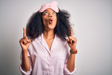 Young african american woman with afro hair wearing sleeping eye mask and pink pajama amazed and...