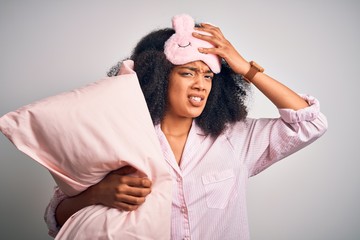 Young african american woman with afro hair wearing pajama and sleep mask holding pillow stressed with hand on head, shocked with shame and surprise face, angry and frustrated. Fear and upset