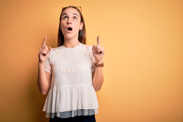 Young beautiful woman wearing casual t-shirt and sunglasses over isolated yellow background amazed and surprised looking up and pointing with fingers and raised arms.