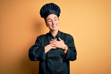 Young beautiful chef woman wearing cooker uniform and hat standing over yellow background smiling with hands on chest with closed eyes and grateful gesture on face. Health concept.