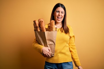 Young beautiful woman holding a bag of fresh healthy bread over yellow background winking looking at the camera with sexy expression, cheerful and happy face.