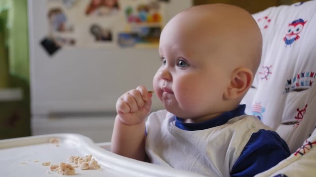 Hungry nine-month-old child sits in a child's chair and eats a pieces of meat.