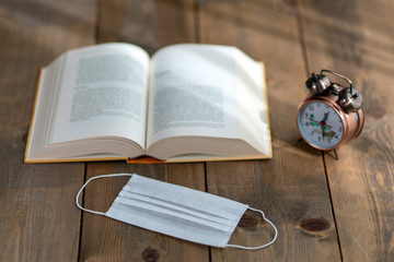 A hardback book opened for reading and a desk clock next to it. Clock book and mask on wooden background