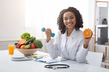 Happy woman doctor holding orange and barbell in clinic