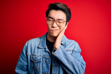 Fototapeta premium Young handsome chinese man wearing denim jacket and glasses over red background touching mouth with hand with painful expression because of toothache or dental illness on teeth. Dentist