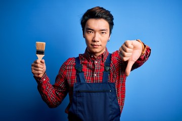 Young handsome chinese painter man wearing apron painting using brush with angry face, negative sign showing dislike with thumbs down, rejection concept