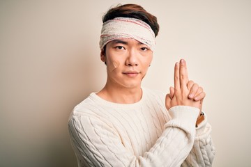 Young handsome chinese man injured for accident wearing bandage and strips on head Holding symbolic gun with hand gesture, playing killing shooting weapons, angry face