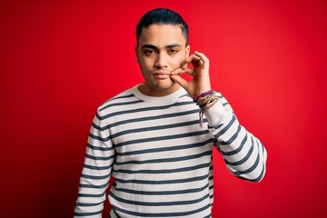 Young brazilian man wearing casual striped t-shirt standing over isolated red background mouth and...