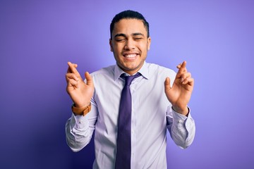 Young brazilian businessman wearing elegant tie standing over isolated purple background gesturing finger crossed smiling with hope and eyes closed. Luck and superstitious concept.