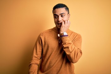 Young brazilian man wearing casual sweater standing over isolated yellow background looking stressed and nervous with hands on mouth biting nails. Anxiety problem.