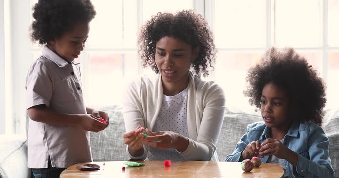 Happy african american children using colorful dough, making different shapes with plasticine with young mother or babysitter at home, creative family kids activity hobby weekend pastime concept.