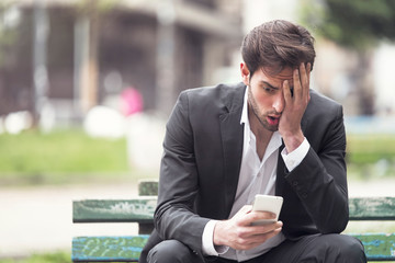 Attractive young businessman sitting on a bench in the park, receiving some disturbing news on his...