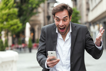 Attractive young businessman holding his phone, reading a message and looking astonished