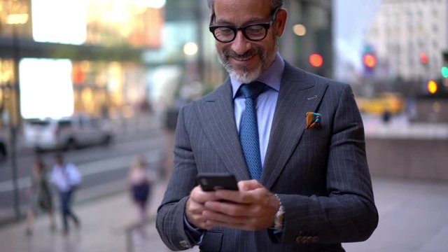 Cheerful middle aged boss using cellular for mobility standing at urban setting in financial district and smiling, happy businessman reading received text message connected to 4g wireless
