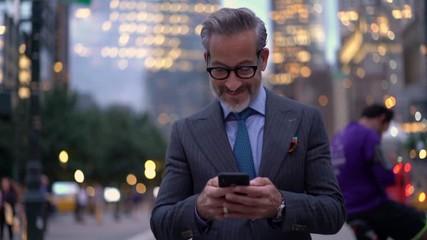 Slow motion of smiling mature lawyer in expensive suit messaging with customers via cellular device while walking in financial district,successful businessman in optical eyewear searching trading news