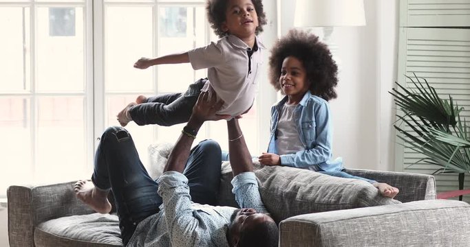 Happy african ethnic kids siblings doing exercises with strong father on comfortable sofa. Smiling mixed race male parent enjoying gymnastics with teen daughter and preschool son in living room.