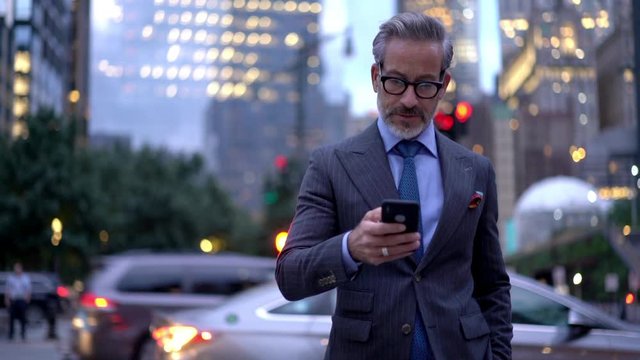 Mature male employer formally dressed walking in metropolitan downtown and typing text message for send via smartphone device, middle aged businessman using mobile device for phoning outdoors
