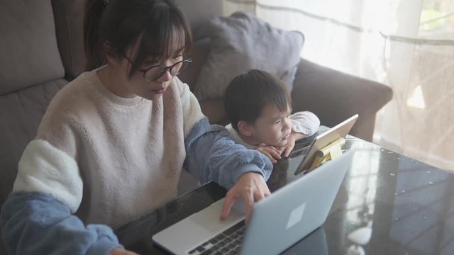 Young asian woman at her home workplace in the living room working with her child watching cartoon on tablet during the coronavirus global  lockdown