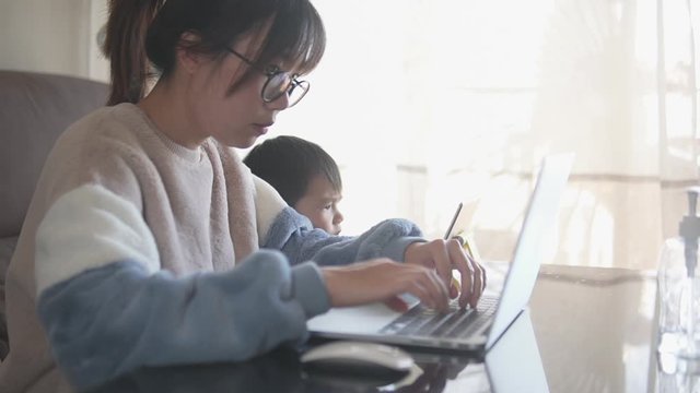 Young asian woman online business owner working on a laptop  from her home office in the living room while taking care of a kid during the coronavirus lockdown stay home pandemic campaign