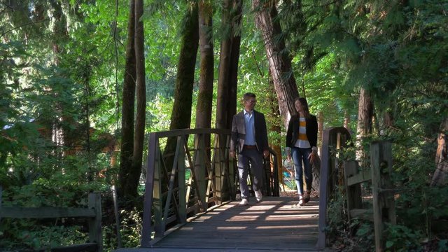 Savvy young, good looking business professionals walk towards camera in a park over a beautiful shaded wooden bridge outside in nature on a sunny day.