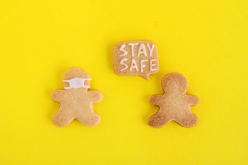 Homemade cookies in shapes of people with inscription ‘Stay safe’ and with face medical mask on yellow background, top view. Sweet shortbread with white glaze. Social distancing concept.