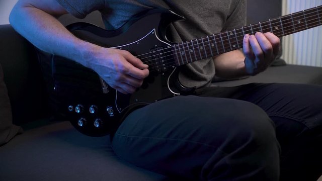 Slider shot of man practicing scales on electric guitar at home sitting on couch.