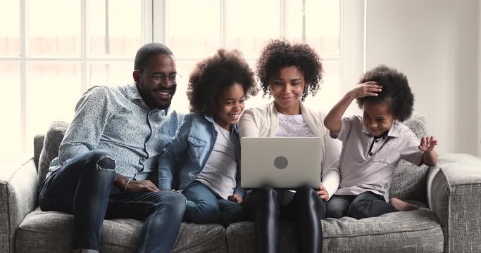Attractive young african ethnicity woman holding on lap computer, watching funny video, shopping in online store or web surfing information with happy husband, teen daughter and preschool son at home.
