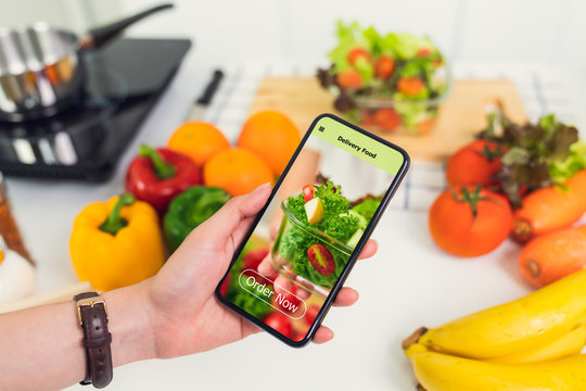 Woman using smartphone and touching application screen for ordering salad online on the table at the kitchen.