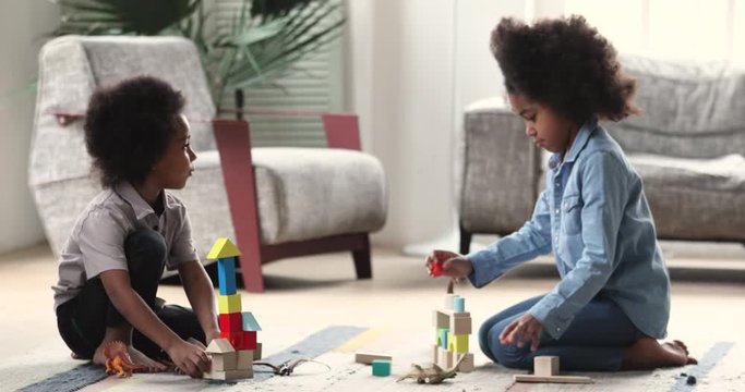 Full length small african american children siblings sitting on warm heated floor, playing favorite toys in living room. Happy little mixed race boy involved in game with older sister at home.