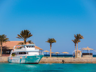 Egyptian beach in february. White ship moored to wharf, medium view. Geen thatch palms under clear blue sky. Selective soft focus. Blurred background