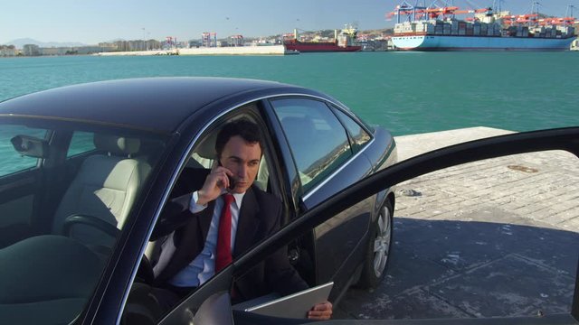 WS Businessman sitting in car on quayside in port, using laptop and phone