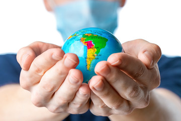 Close-up of a man's hands holding a small globe, with images South America: Brazil, Argentina,   a man in a medical mask out of focus, dressed in a  t-shirt, uniform on a white background. Coronavirus