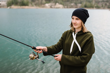 Woman catching a fish, pulling rod while fishing. Girl fishing from pier on lake or pond with text space. Fisherman with rod, spinning reel on river bank. Fishing for pike, perch, carp. Wild nature.