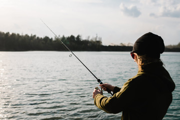 Fisherman with rod, spinning reel on the river bank. Sunset. Fishing for pike, perch, carp. Woman catching a fish, pulling rod while fishing. Girl fishing from beach lake or pond with text space.