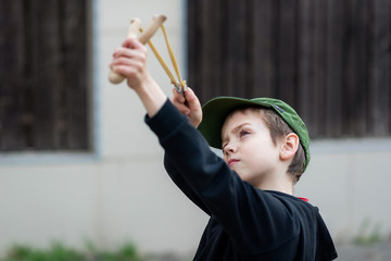 A boy in a black jacket with a hood and a cap shoots from a slingshot, blurred background