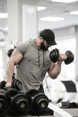 Fototapeta na wymiar strong young caucasian male with beard in cap and sportswear lifting heavy dumbbell indoors fitness gym listening music for motivation