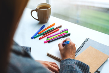 Closeup image of a woman writing on a blank notebook with colored pens and coffee cup on the table