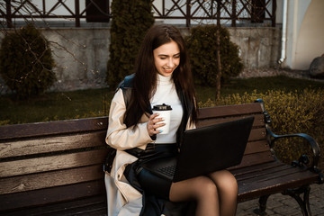 close portrait of a girl with a laptop sitting on a bench with a cup of coffee. Student with laptop studying in the street.