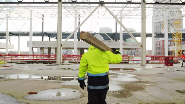 Rear view of man worker walking outdoors on construction site, carrying beam.