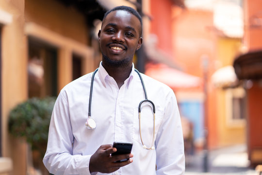 African Doctor With Stethoscope Holding A Smartphone Near Modern Clinic Hospital Building