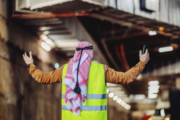 Rear view of arabian investor in vest, with headscarf holding smart phone and standing with opened...